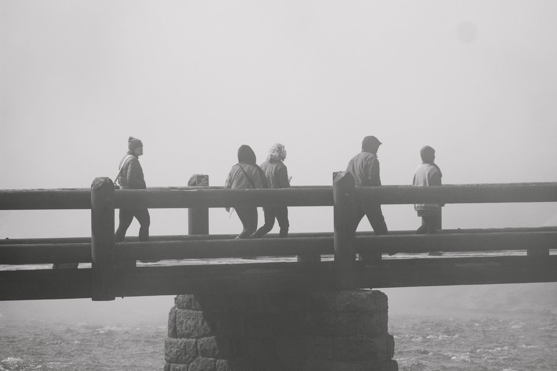 Black and white photograph of 5 figures crossing a bridge over water and fog.
